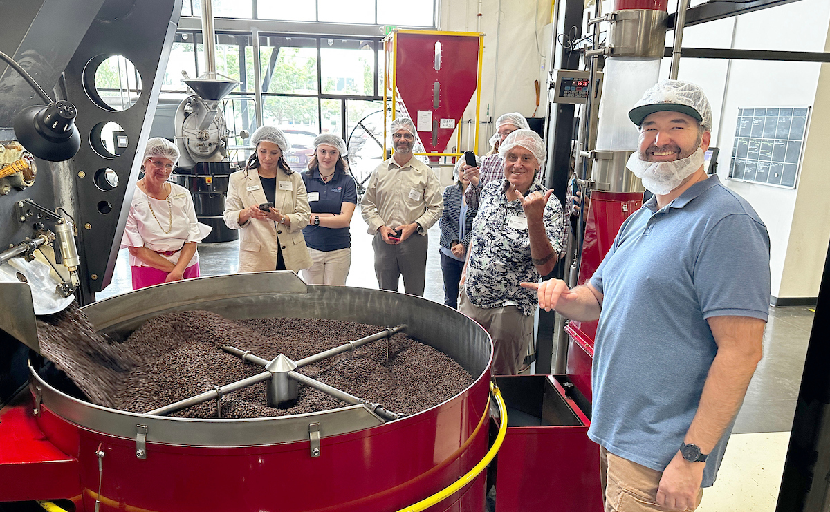 A man stands near an industrial coffee grinder with a group of onlookers in hair nets.